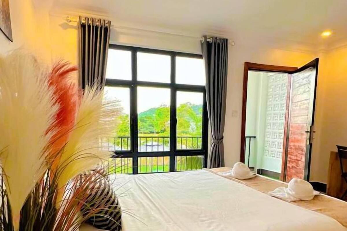 A hotel room with a large bed, a view of greenery outside, and a wooden side table with a vase of flowers.