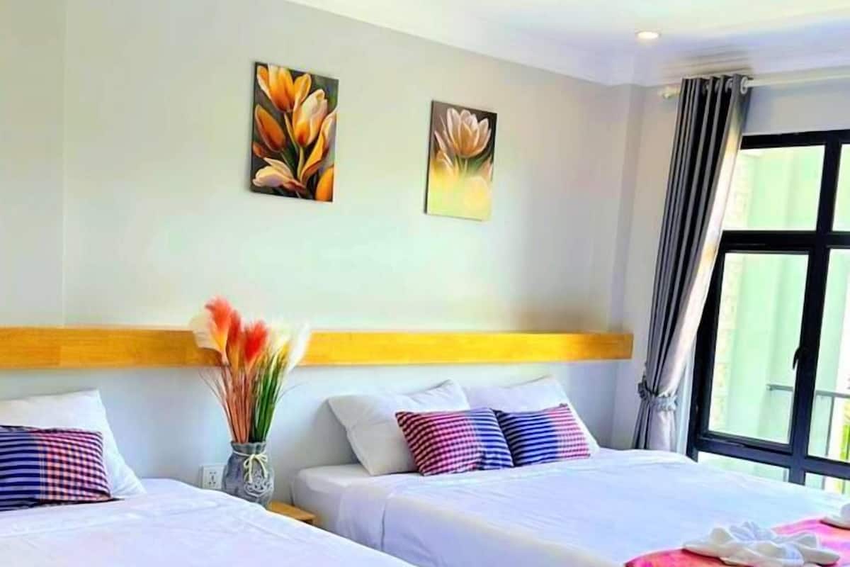A hotel room with two beds, each with a white bedspread and a red runner. There are floral paintings on the wall, a vase with flowers, and a window with curtains.