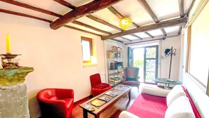 Een kamer met zichtbare houten balken, een rode fauteuil, een witte bank, een houten salontafel en een boekenplank.