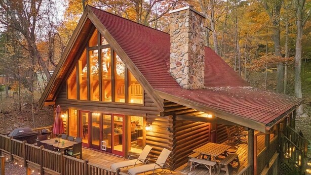 A log cabin with a stone chimney, a deck with outdoor seating, and a large window overlooking the forest.