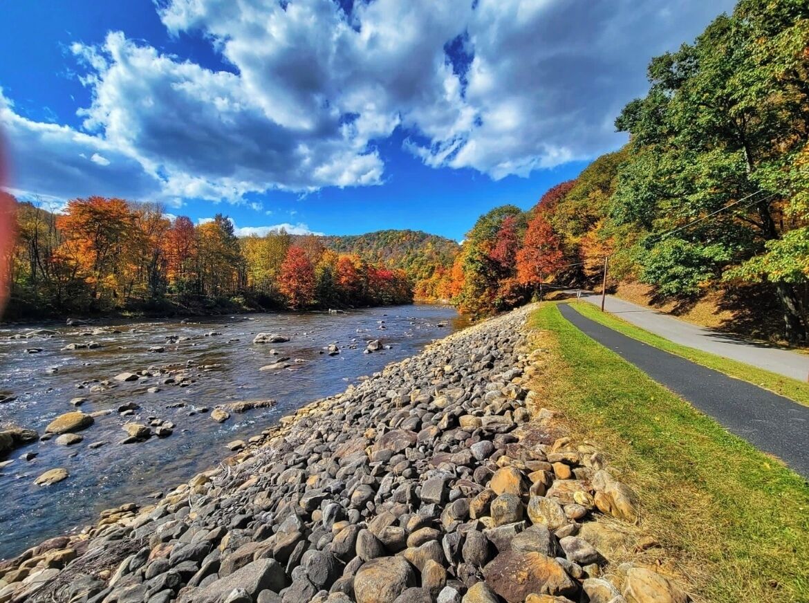 A river with a rocky shore, a paved path alongside, and trees with autumn foliage in the background.