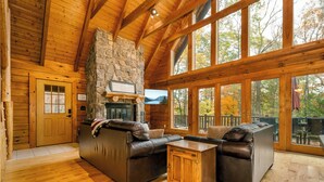 A log cabin interior with a stone fireplace, leather sofas, and large windows offering a view of trees.