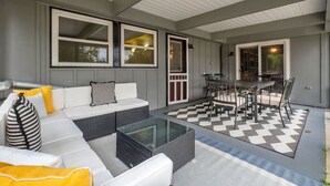 Een overdekt terras met een witte bank, een glazen salontafel en een eettafel met stoelen.
