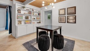 Een modern interieur met een houten plafond, witte muren, een zwarte tafel met poefs en planken met boeken en gezelschapsspellen.