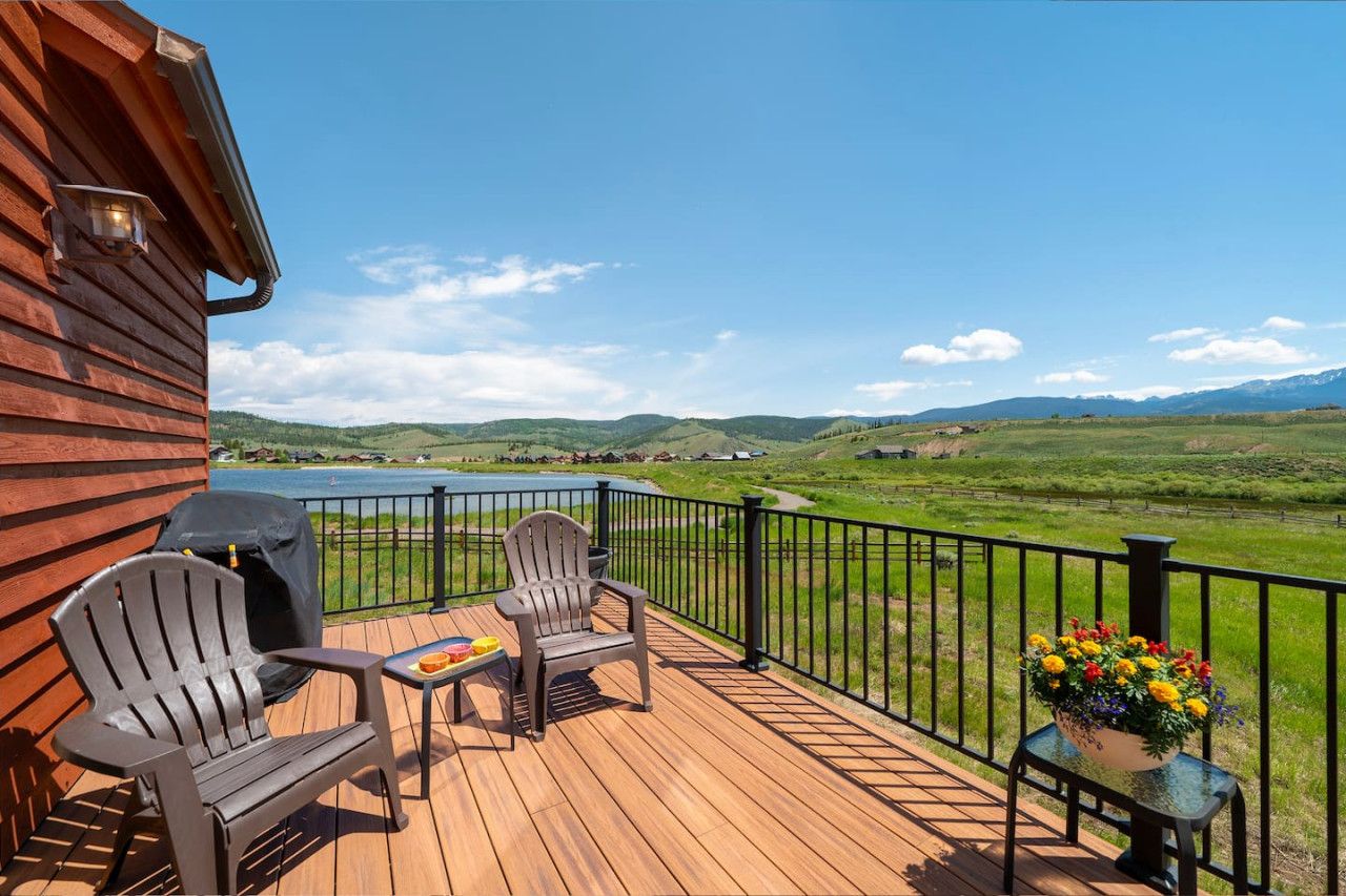 Eine Holzterrasse mit zwei Adirondack-Stühlen, einem Grill und einem Tisch mit Buch und Snacks. Ein Blumentopf steht am Terrassenrand.