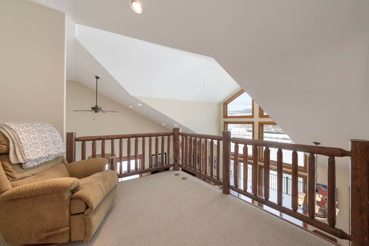 A spacious loft bedroom with a high ceiling, a wooden railing, a ceiling fan, and a large window offering a view of the outdoors.