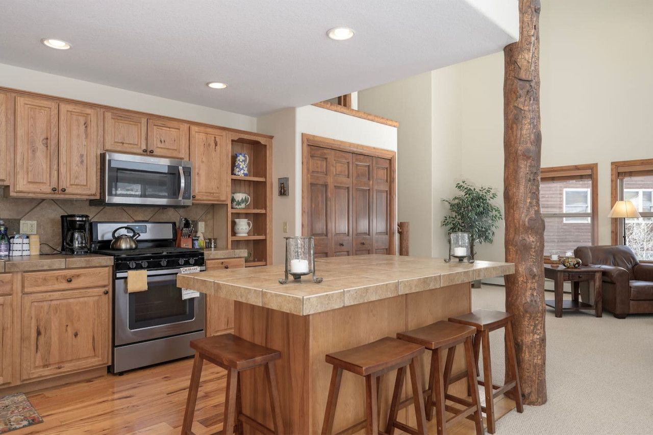 A kitchen with wooden cabinets, a stainless steel microwave, and a built-in oven. There is a large island with stools and a wooden tree trunk in the center.