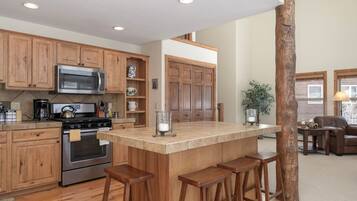 A kitchen with wooden cabinets, a stainless steel microwave, and a built-in oven. There is a large island with stools and a wooden tree trunk in the center.