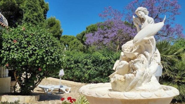 A classical stone fountain with a statue of a woman and child, surrounded by lush greenery and flowering trees. - Oasis on the Montgo, cozy apartment with large covered terrace (Dénia)