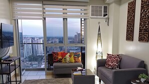 Ein modernes Wohnzimmer mit einer Couch, einem Couchtisch und einem Fernseher. Ein großes Fenster bietet Blick auf die Stadt.