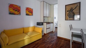 Un espacio habitable compacto con un sofá amarillo, una mesa de comedor con sillas, una zona de cocina y obras de arte en las paredes.