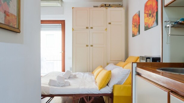 Habitación de hotel compacta con cama, kitchenette pequeña, armario y ventana con persianas.