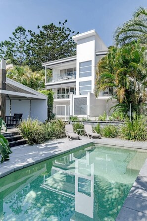 A modern two-story house with a swimming pool, outdoor seating, and lush greenery.