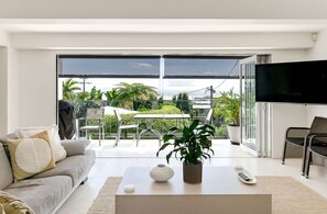A modern living room with a sofa, coffee table, and TV. There is a balcony with outdoor furniture and a view of greenery.