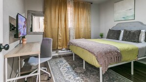 A bedroom with a bed, a desk, a chair, a ceiling fan, and a window with curtains.