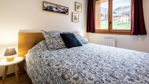 Ein Schlafzimmer mit Bett, Nachttisch, Lampe, einem Fenster mit Vorhängen und gerahmten Bildern an der Wand.