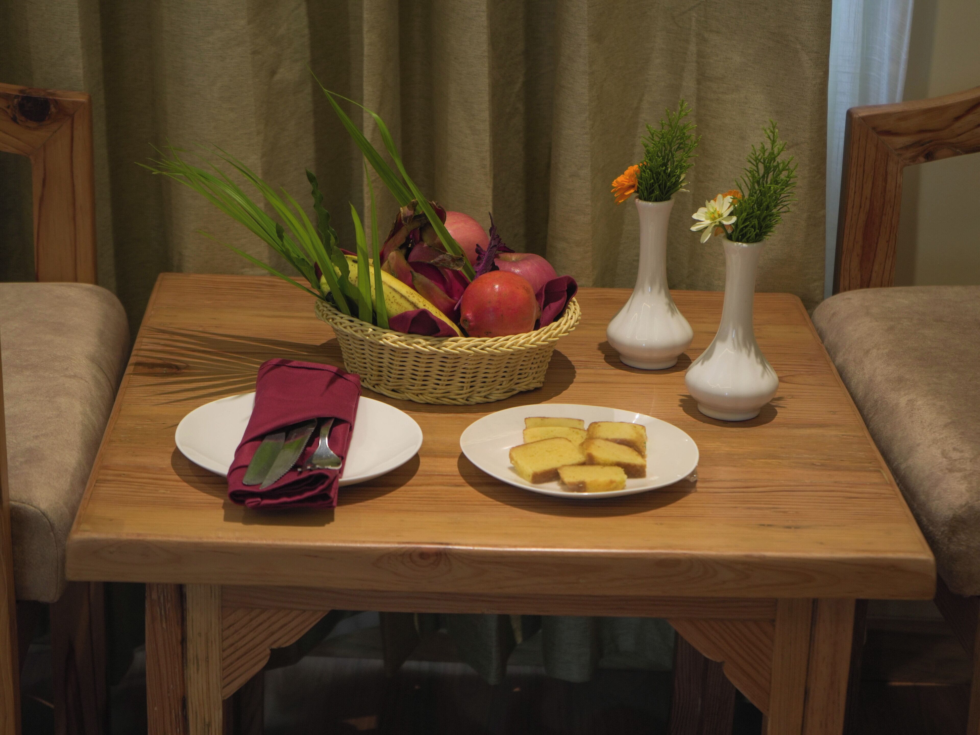 A wooden table with a basket of fruits, two vases with flowers, and plates with food.
