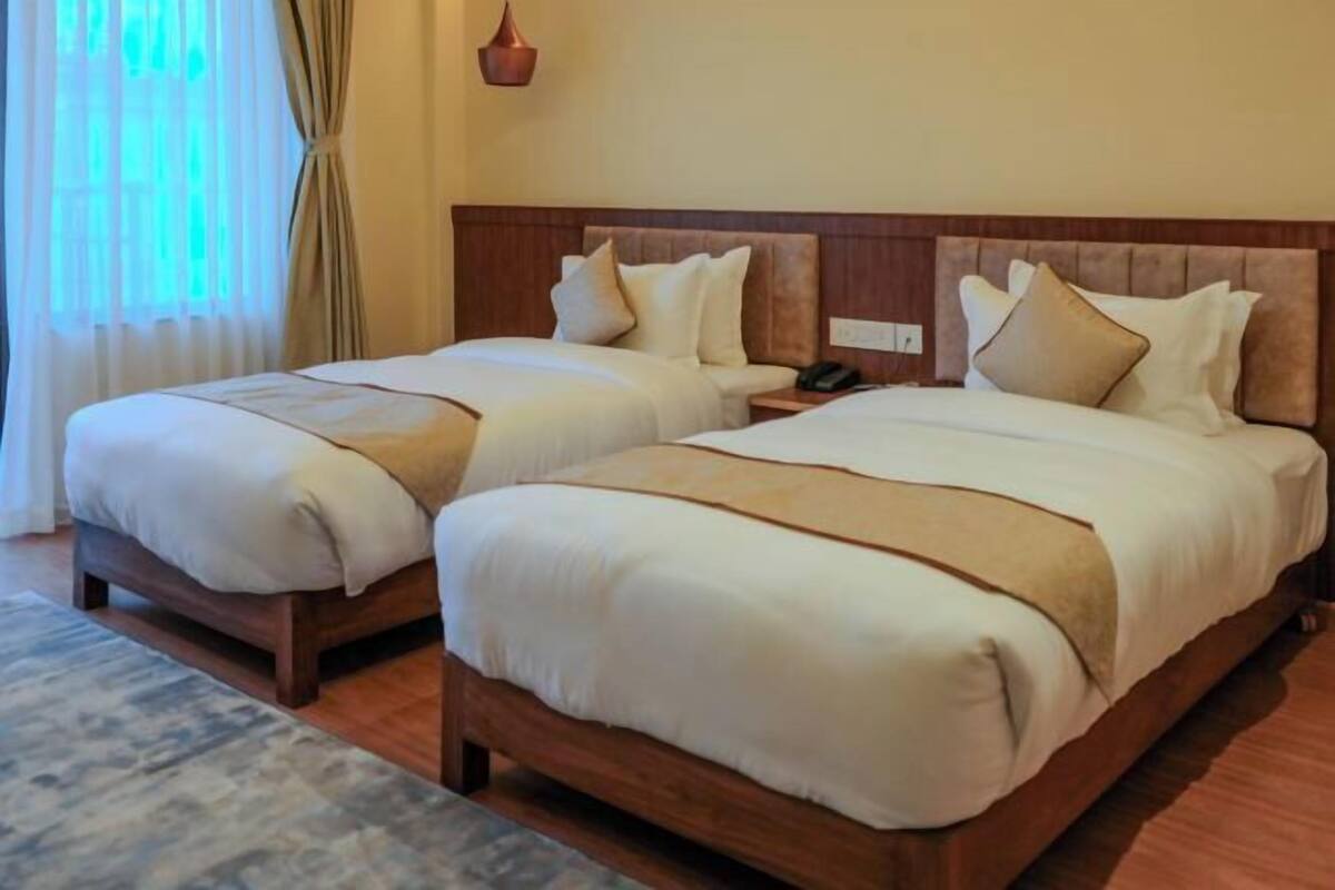 A hotel room with two beds, wooden headboards, beige bedding, a bedside table with a phone, and a window with curtains.