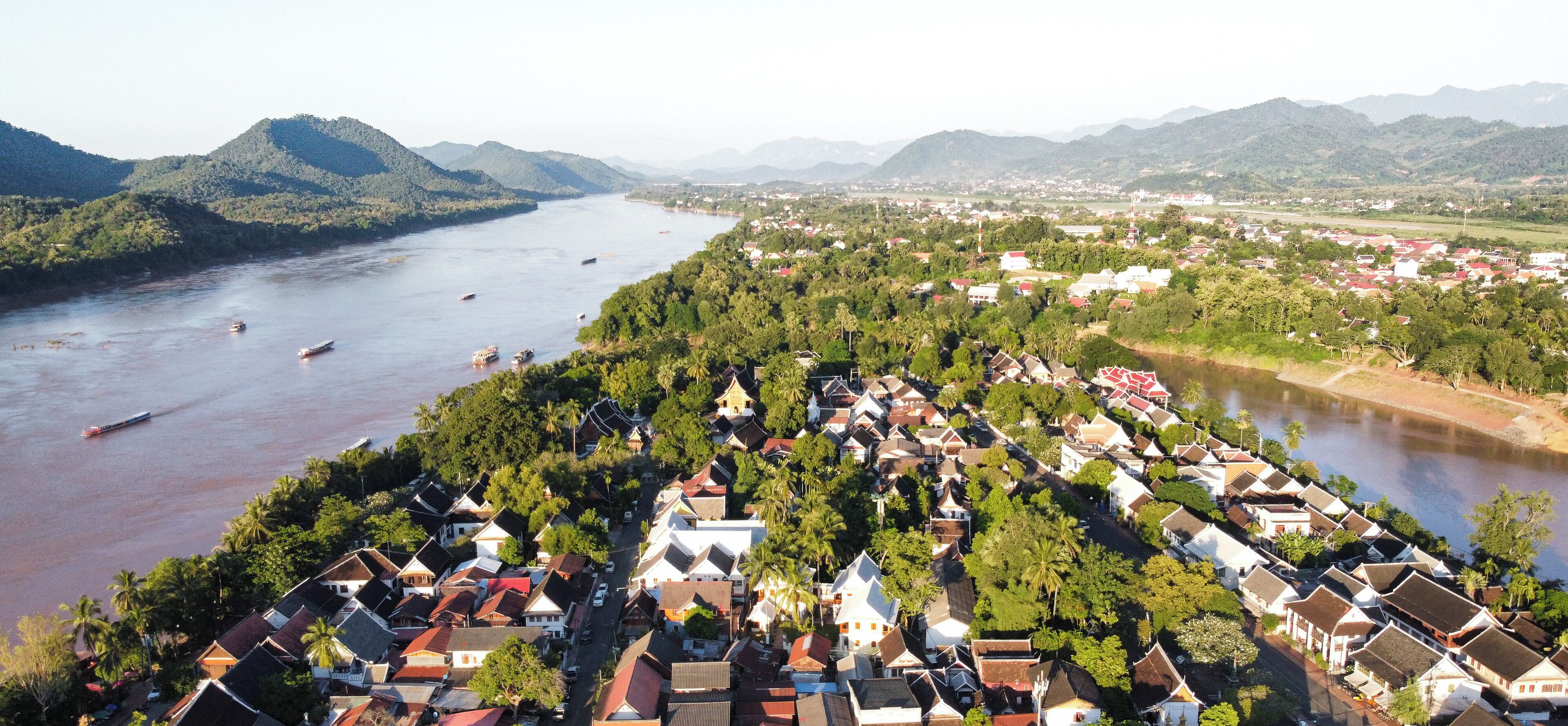 Uma cidade às margens do rio, com casas tradicionais, o rio e montanhas ao fundo.