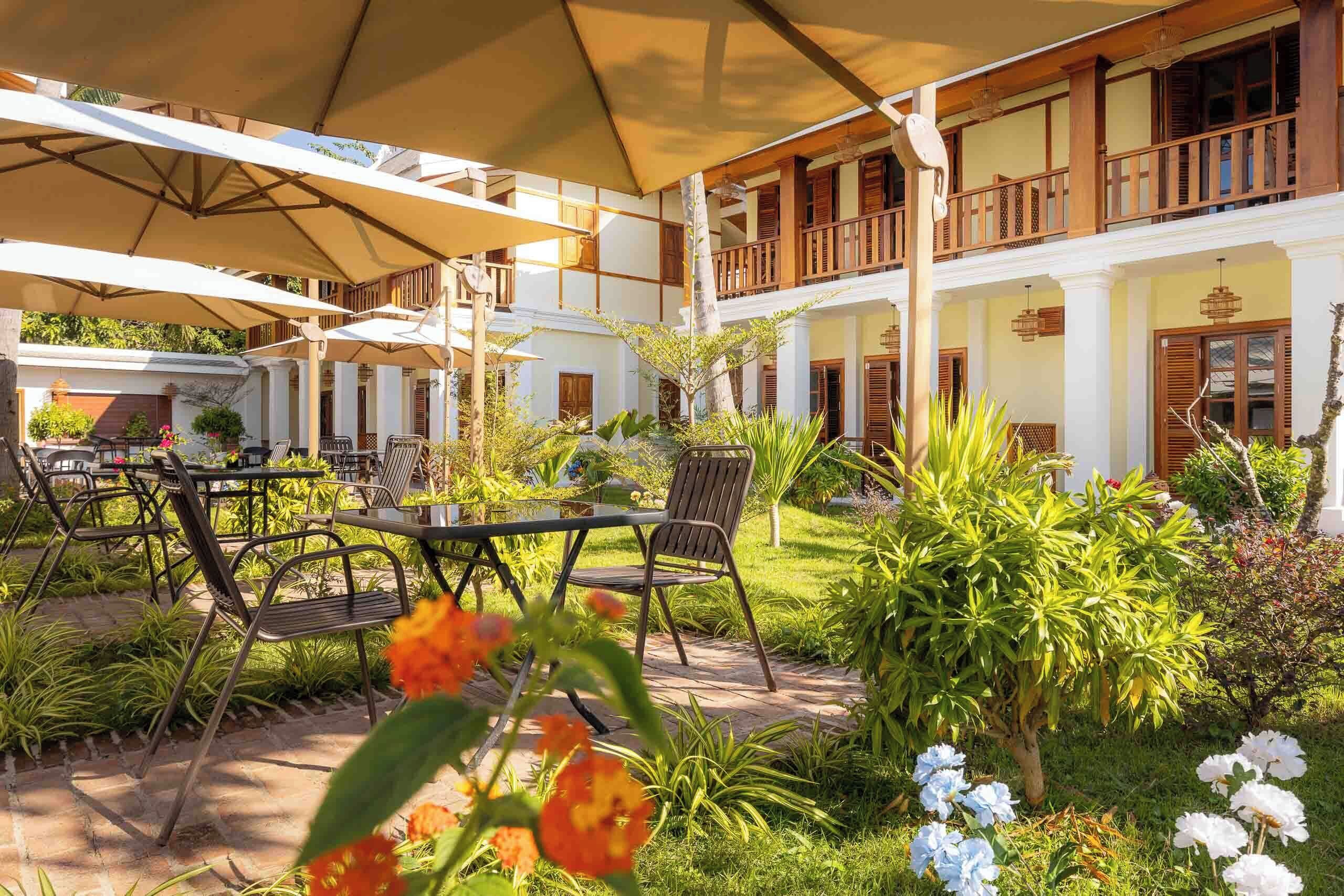 Un patio con mobiliario exterior, sombrillas y un jardín con diversas plantas y flores.