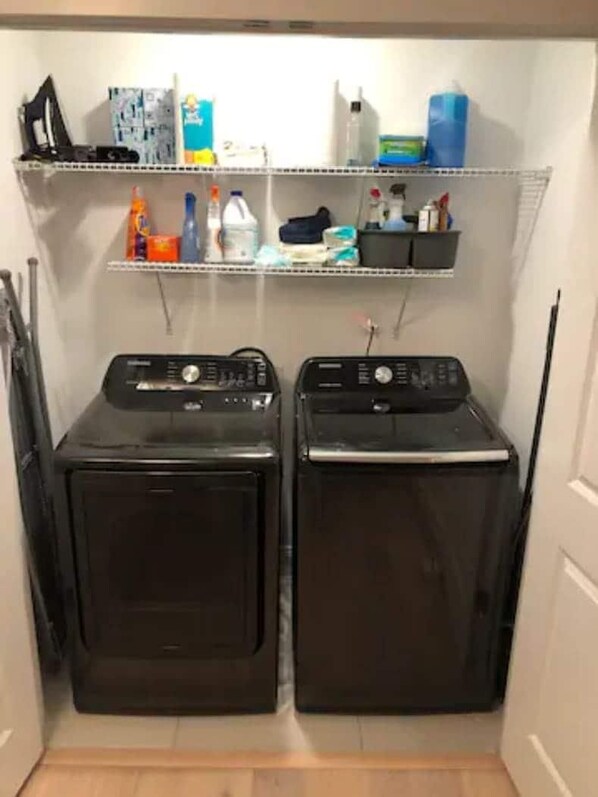 A laundry room with two front-loading washing machines and a shelf above containing various cleaning supplies.
