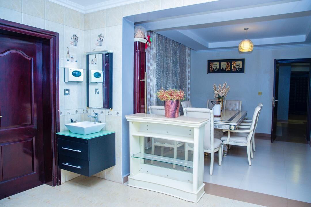 A modern bathroom with a dark wooden door, a white sink, and a vanity with drawers. There is a mirror above the sink and a vase with flowers on a table. The room has a blue accent wall and a dining area with a table and chairs.
