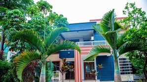 A modern building with a blue and red facade, surrounded by lush greenery and palm trees.