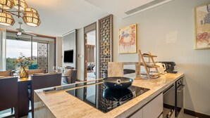 A modern kitchen with a glass cooktop, a dining area with a table and chairs, and a view of the outdoors through large windows. - Angsana Oceanview Residences by Banyan Living (Choeng Thale)