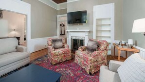 Un salon doté d’une cheminée, de deux fauteuils à motifs, d’un canapé, d’un téléviseur fixé au-dessus de la cheminée et d’une petite table avec des lampes.