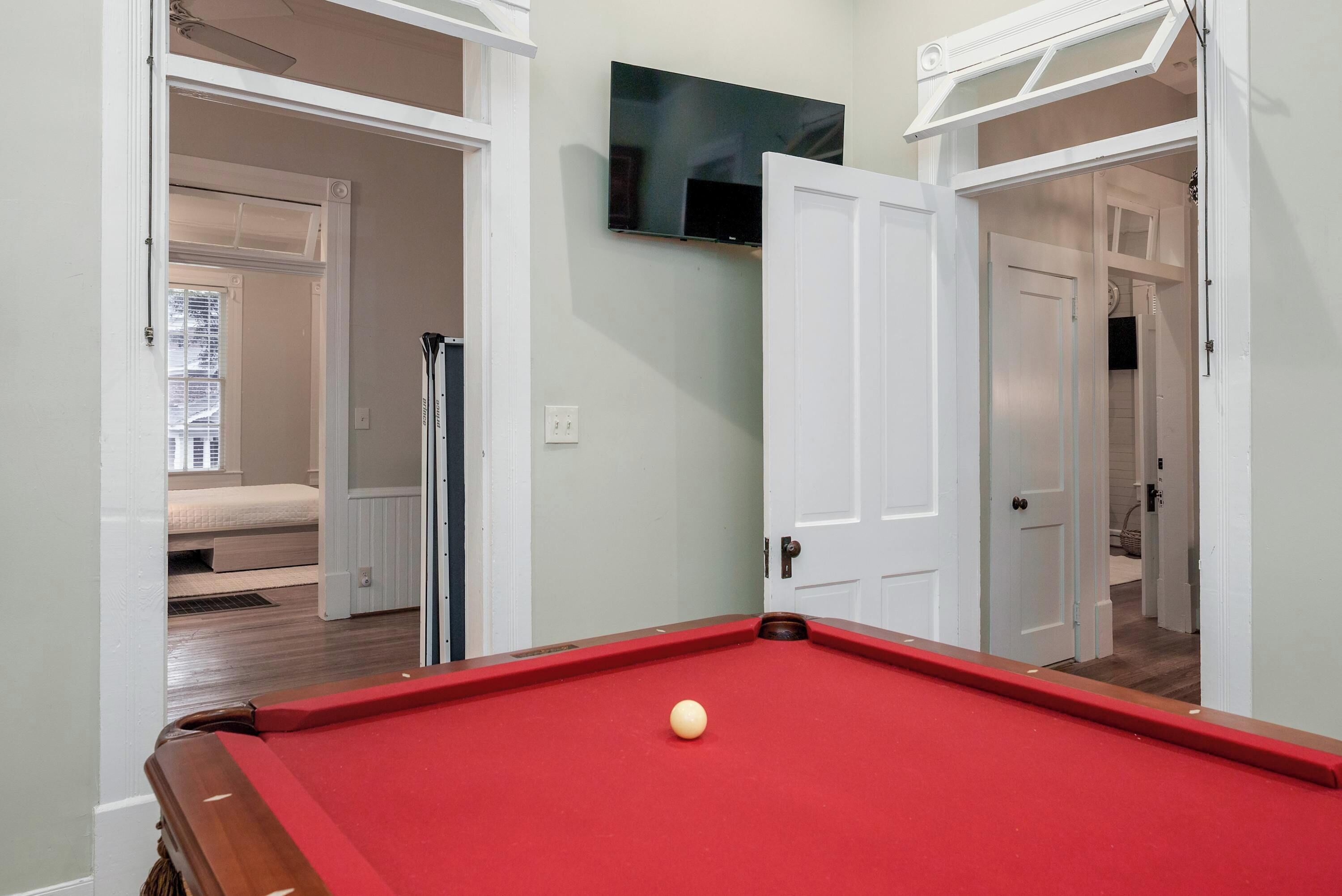 A pool table with a single ball, a television mounted on the wall, and open doors leading to other rooms.