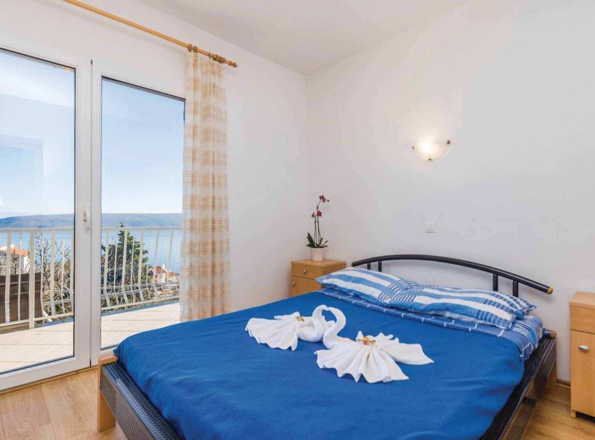 A bedroom with a large bed, a nightstand, a potted plant, and a view of the sea through a sliding glass door.