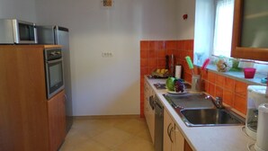 Et kompakt køkken med mikrobølgeovn, ovn og vask. Der er orange fliser, et vindue med gardin og en lille plante på bordet.
