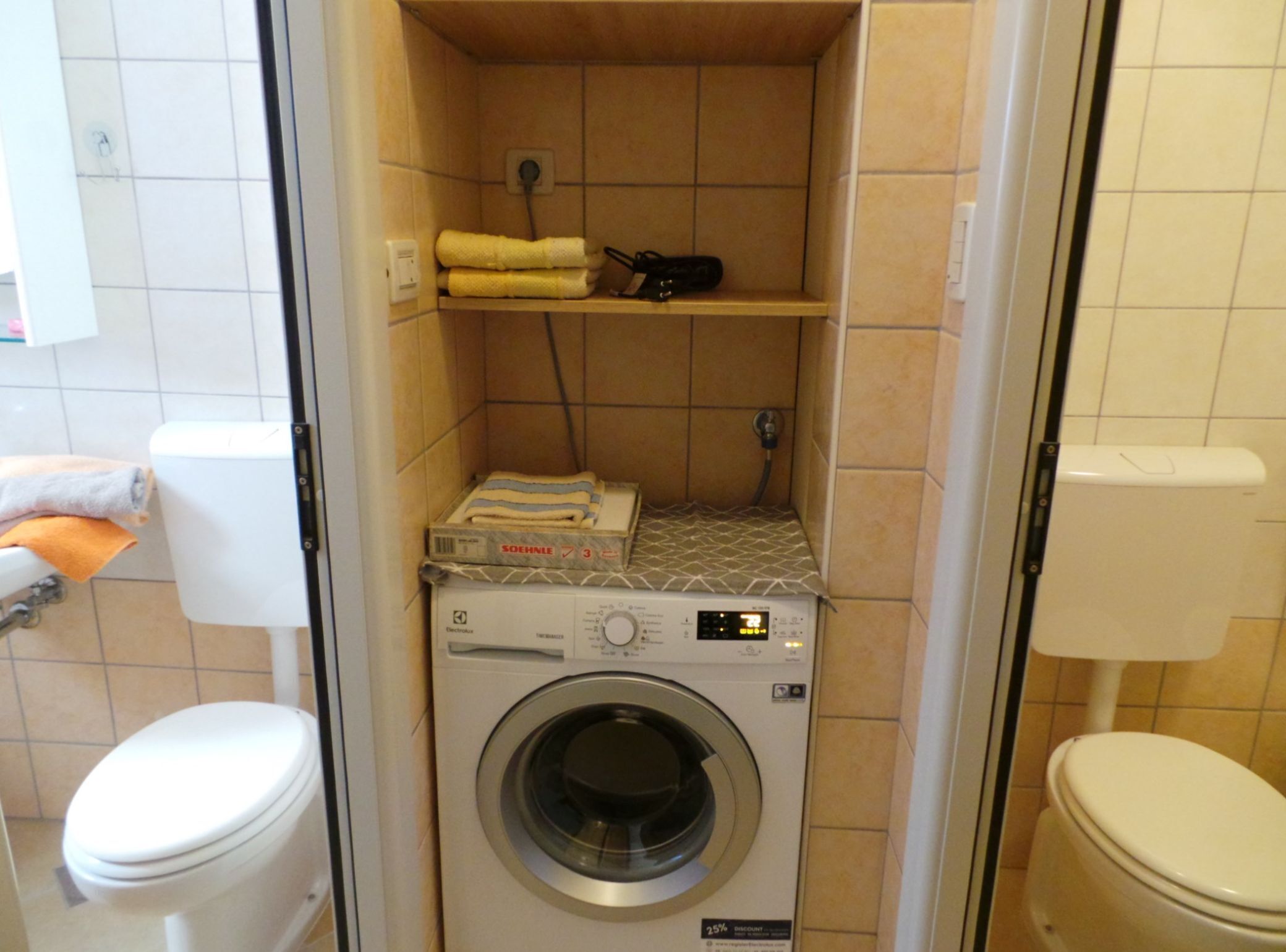 A compact bathroom with a washing machine, a toilet, and a shelf with towels.