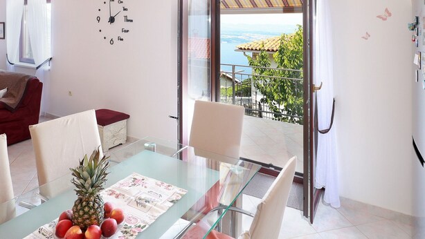 A dining area with a glass table, chairs, and a fruit centerpiece. A clock is on the wall, and there's a view of the sea through an open door. - Sunny Holiday Home With Terrace (Lovran)