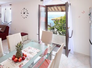 Een eetgedeelte met een glazen tafel, stoelen en een fruitstuk. Een klok hangt aan de muur, en er is uitzicht op de zee door een geopende deur.