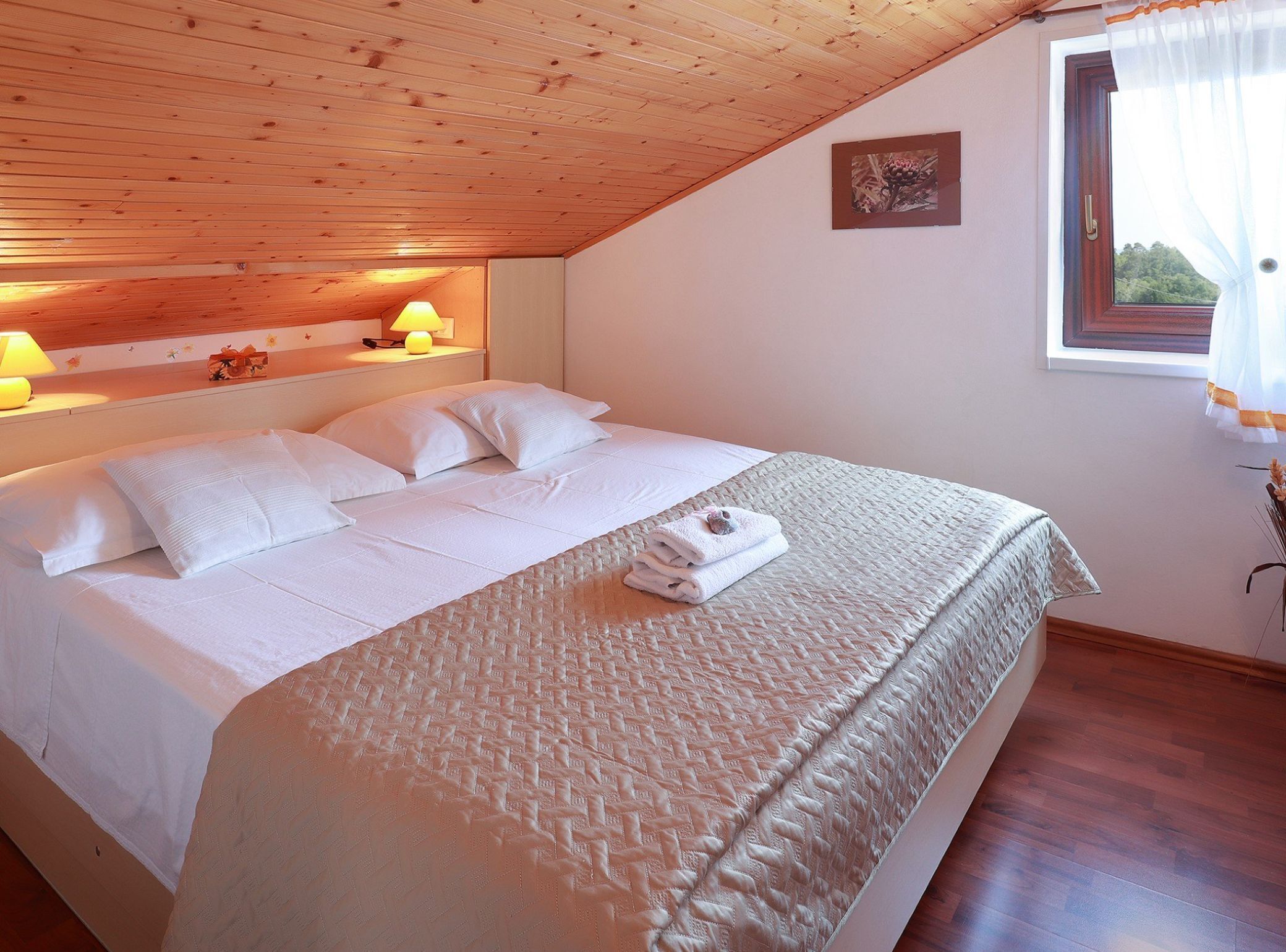 A neatly made bed with white linens, a wooden ceiling, and a window with curtains.