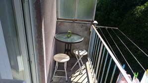 Un balcón con una pequeña mesa, dos taburetes, una barandilla metálica y una ventana.