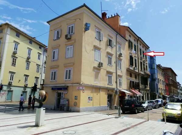 A multi-story building with a yellow facade, multiple windows, and a street in front with parked cars and pedestrians. - Stylish Rijeka City Center Apartment (Rijeka)