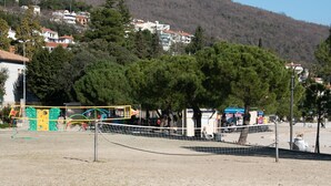 A sandy beach area with playground equipment, trees, and a view of houses in the background. - Cozy Icici Apartment With Sea View (Opatija)