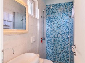 A bathroom with a white toilet, a shower with blue mosaic tiles, and a mirror with a wooden frame. - Beachside Serenity Apartments in Barbot (Rab)