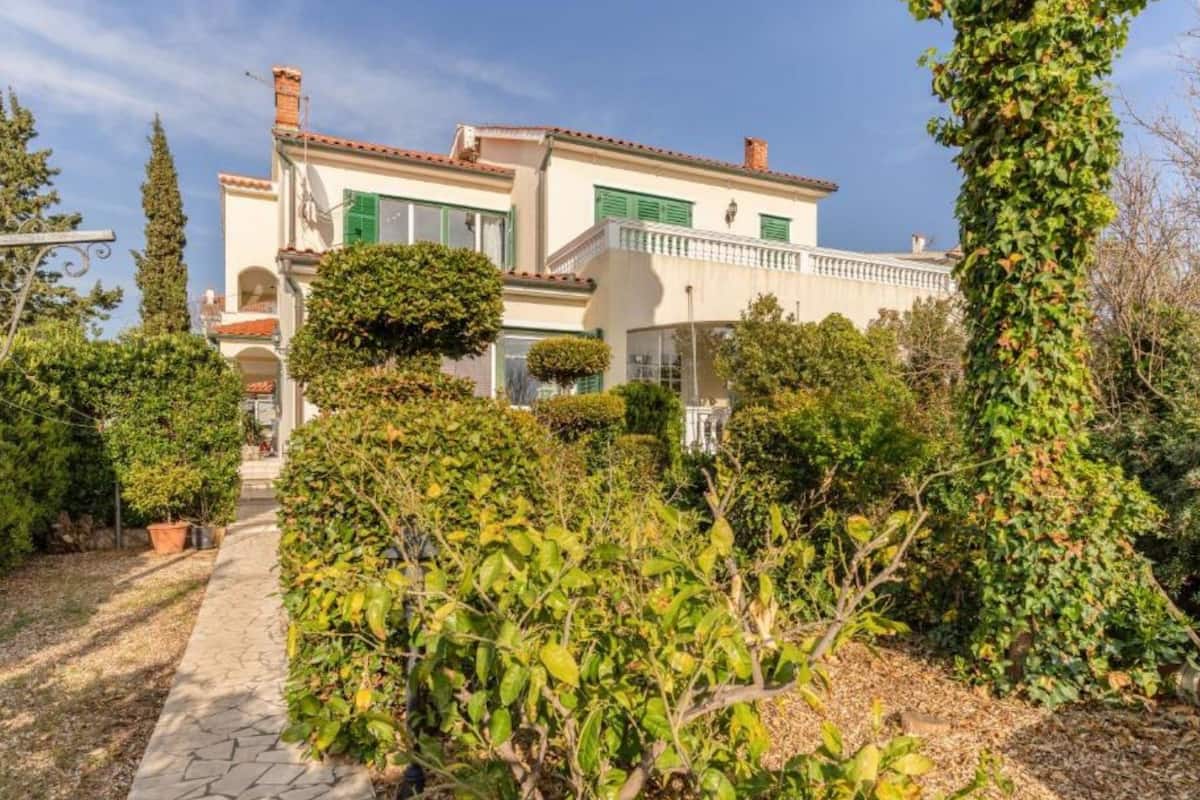 A two-story house with a paved pathway leading to the entrance, surrounded by lush greenery and a variety of plants.