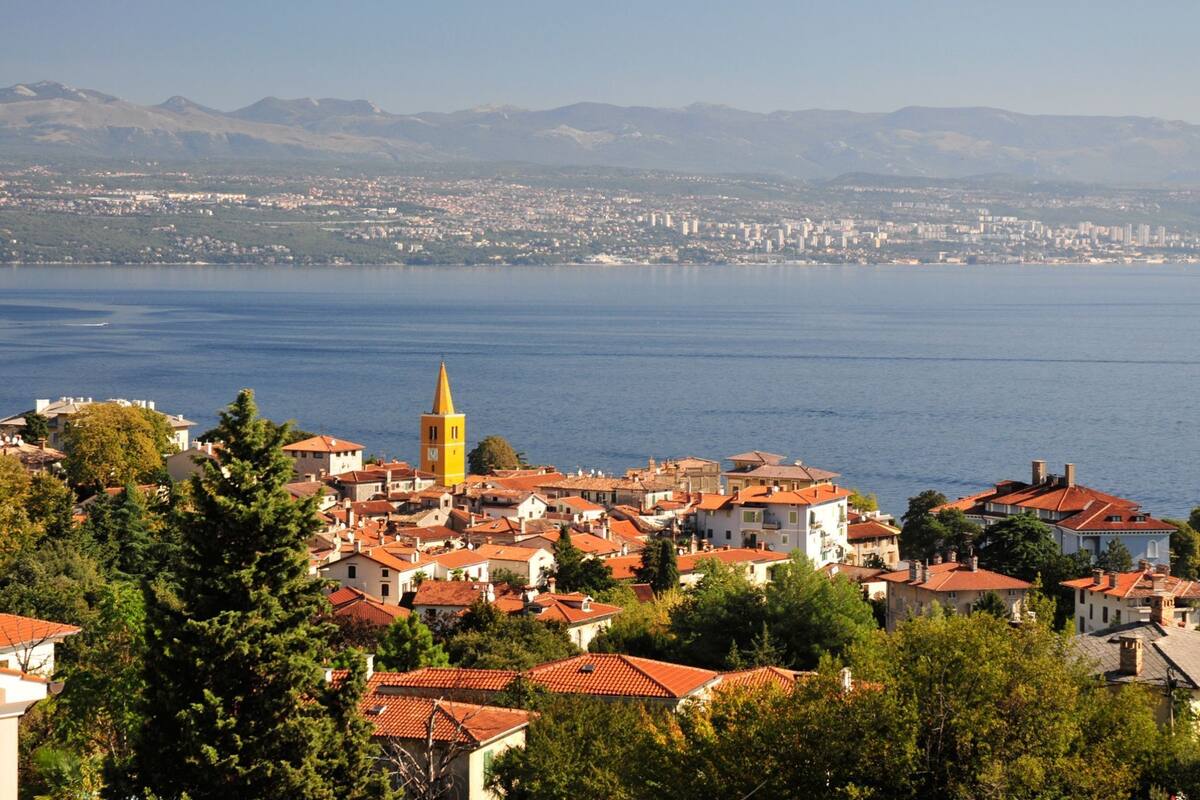 A coastal town with a prominent yellow tower, red-roofed buildings, and a view of the sea and distant mountains.