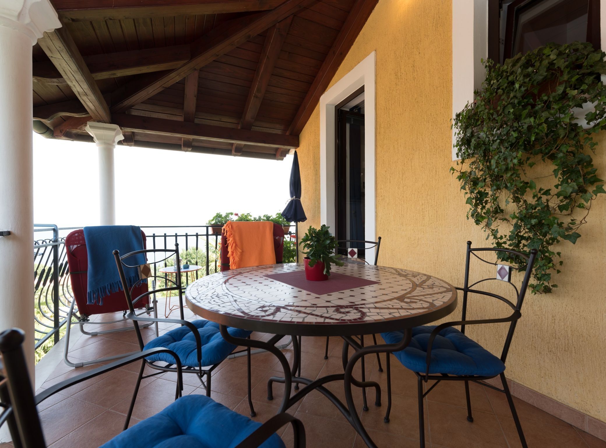 Portico esterno coperto con tavolo rotondo, sedie imbottite blu e una pianta in vaso.