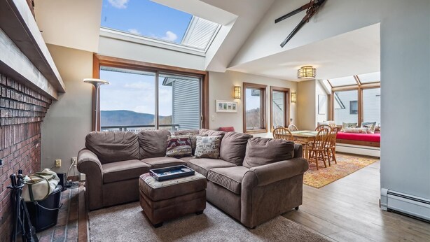 Een ruime woonkamer met een grote hoekbank, een klein voetenbankje en een eethoek met een rode bank en houten stoelen. Een dakraam zorgt voor natuurlijk licht, en de kamer heeft een accentmuur van baksteen en een groot raam.
