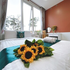 A hotel room with a bed, a window with curtains, a bedside table with a lamp, a mirror, and a vase with flowers.