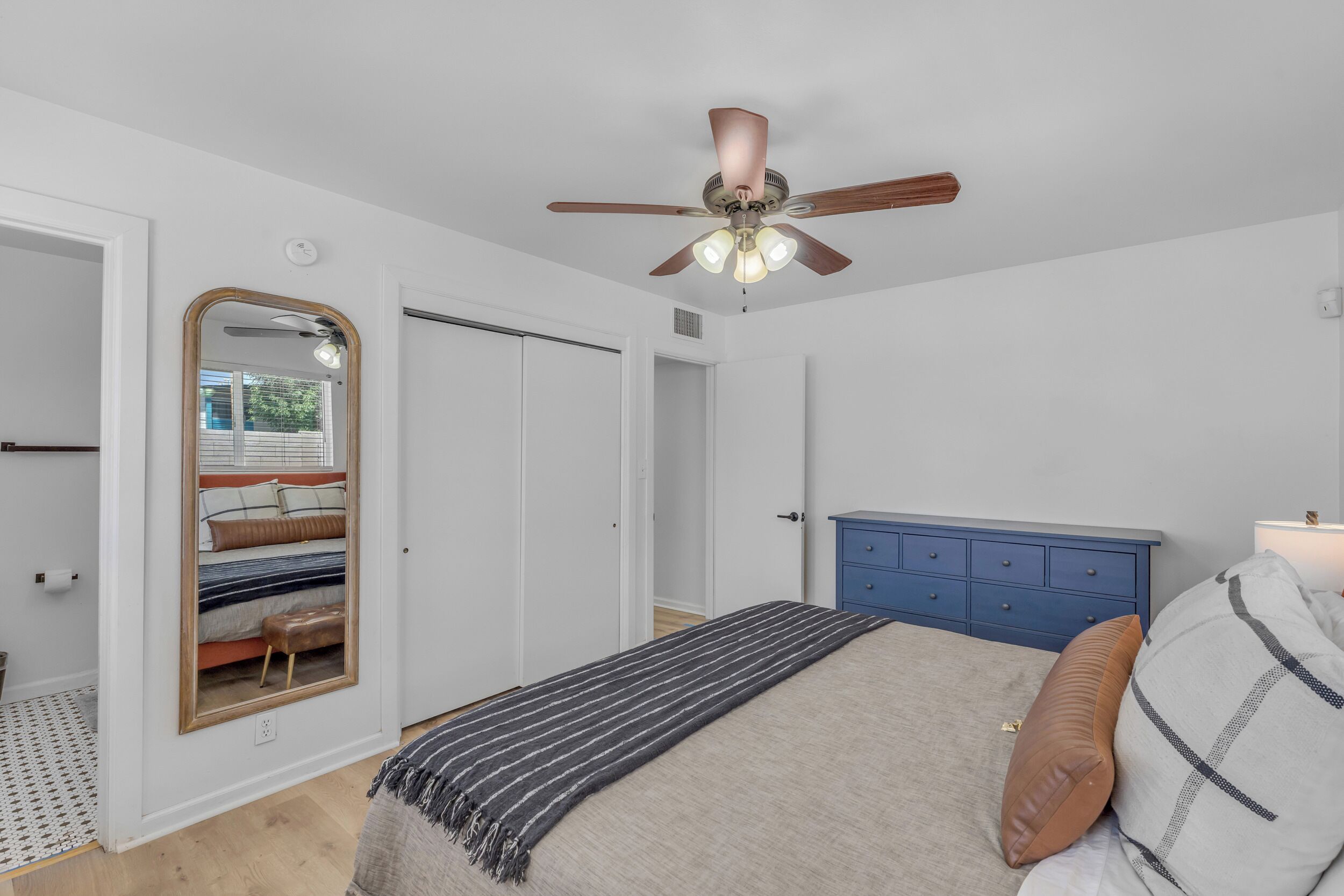 Een slaapkamer met een bed, een blauwe dressoir, een plafondventilator en een spiegel.