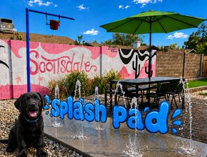 A dog in front of a wall with graffiti, a table and chairs, and a green umbrella.