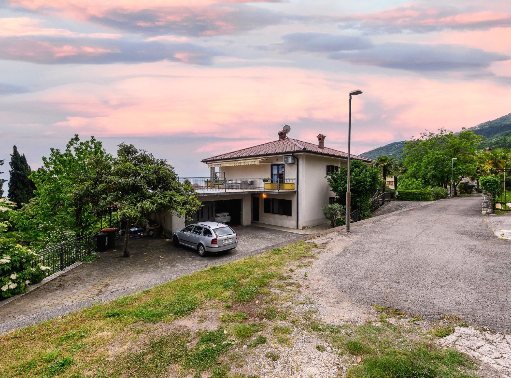 Une maison à deux étages avec une voiture garée dans l’allée, entourée de verdure et avec une montagne en arrière-plan.