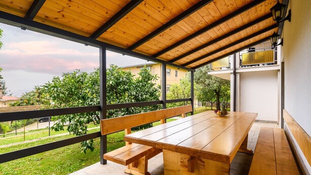 A covered outdoor patio with a wooden table and benches, overlooking a garden and buildings. - Charming Lovran Apartments With Wonderful Sea View (Lovran)