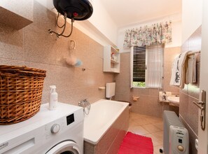 A bathroom with a washing machine, a bathtub, a radiator, and a window with curtains. - Elegant Lovran Apartment With Large Outdoor Area (Lovran)